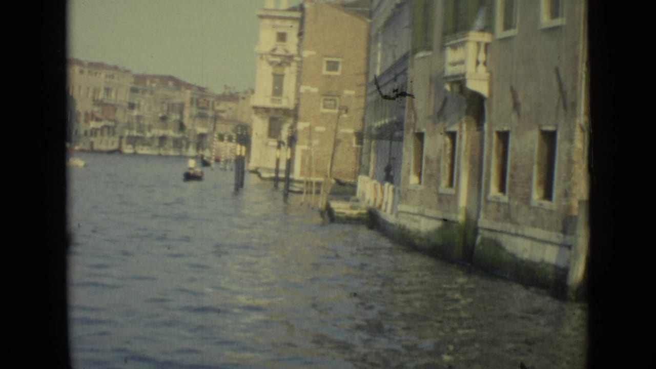 1962 Venice canal footage from a gondola or boat, passing close to aged waterfront walls, windows, mooring posts, and distant boats.
