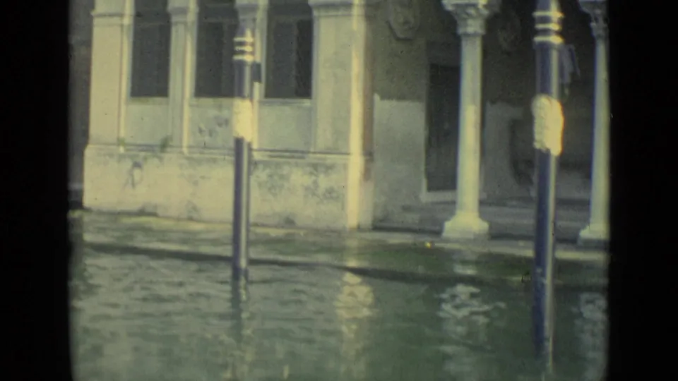 1962 Venice frame with green floodwater, a lamppost, pale columns, and weathered building details along the canal.
