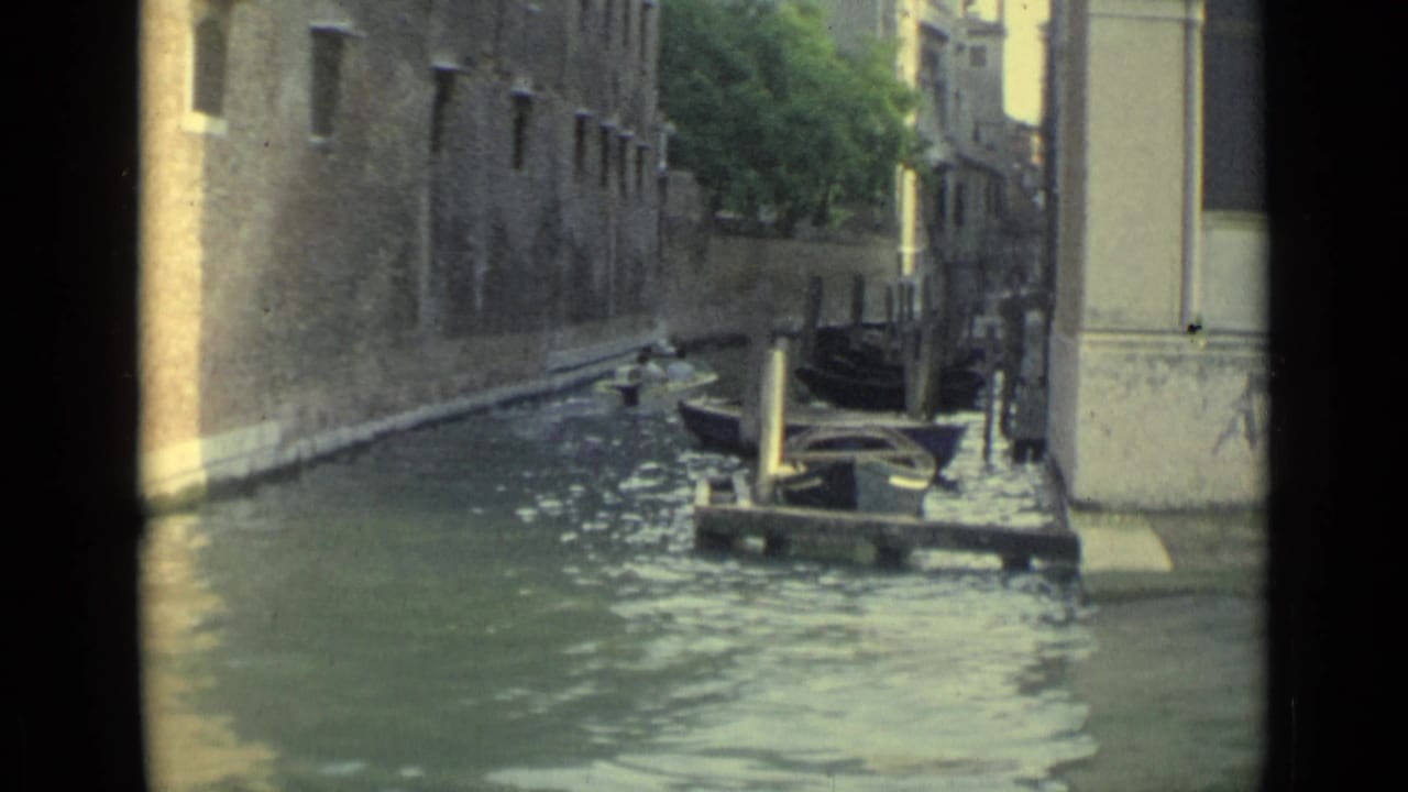 1962 Venice canal scene with a gondola-like boat, moored craft, posts, and sunlit stone walls along the water.