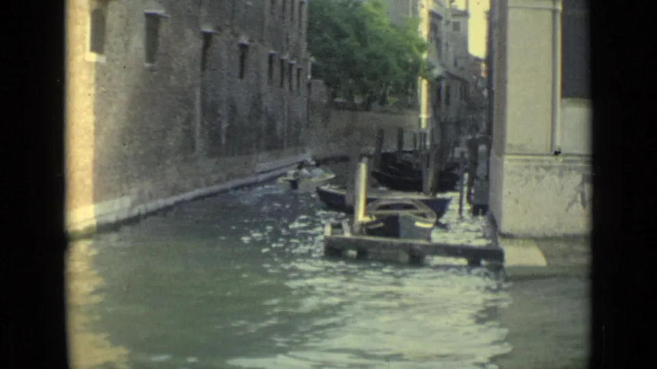 1962 Venice canal scene with a gondola-like boat, moored craft, posts, and sunlit stone walls along the water.