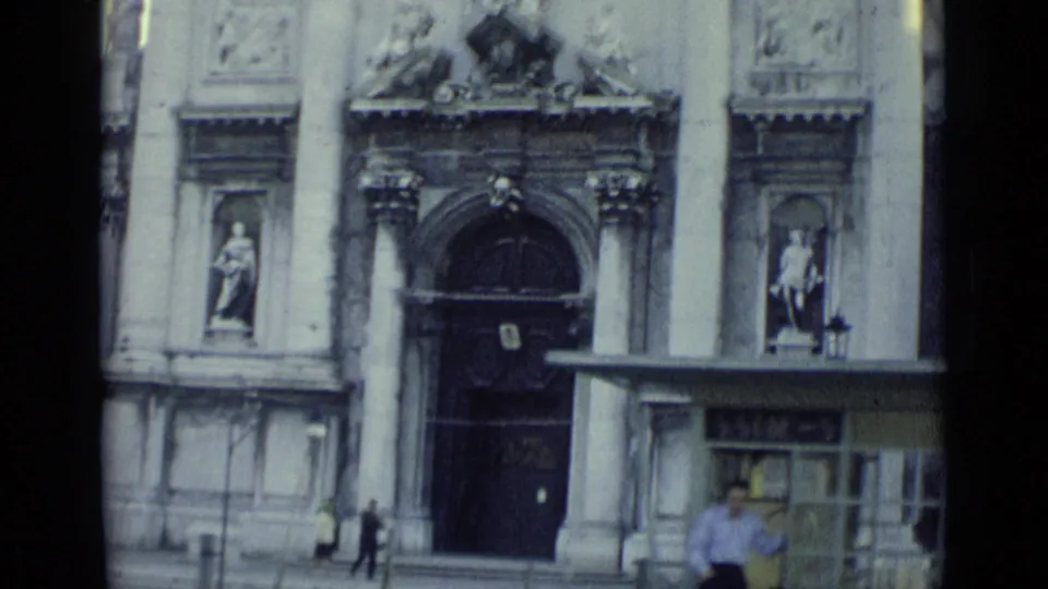 1962 Venice church entrance with statues, columns, a kiosk or shelter, and a few distant pedestrians near the doorway.