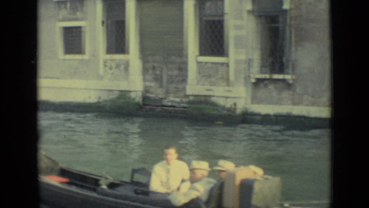 Gondola passengers in light clothing ride through a Venice canal beside aged walls, windows, and a low waterline.