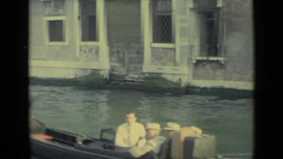 Gondola passengers in light clothing ride through a Venice canal beside aged walls, windows, and a low waterline.