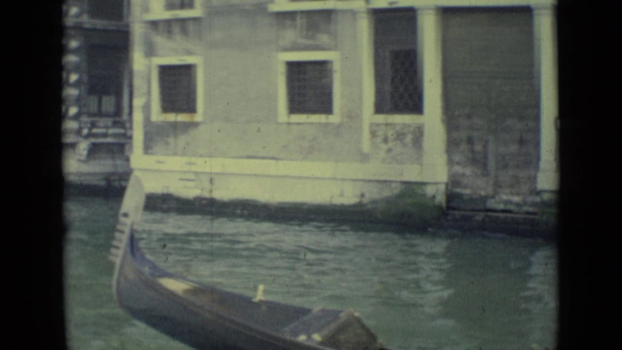 The curved prow of a Venice gondola moves through green canal water beneath shuttered windows and a weathered doorway.