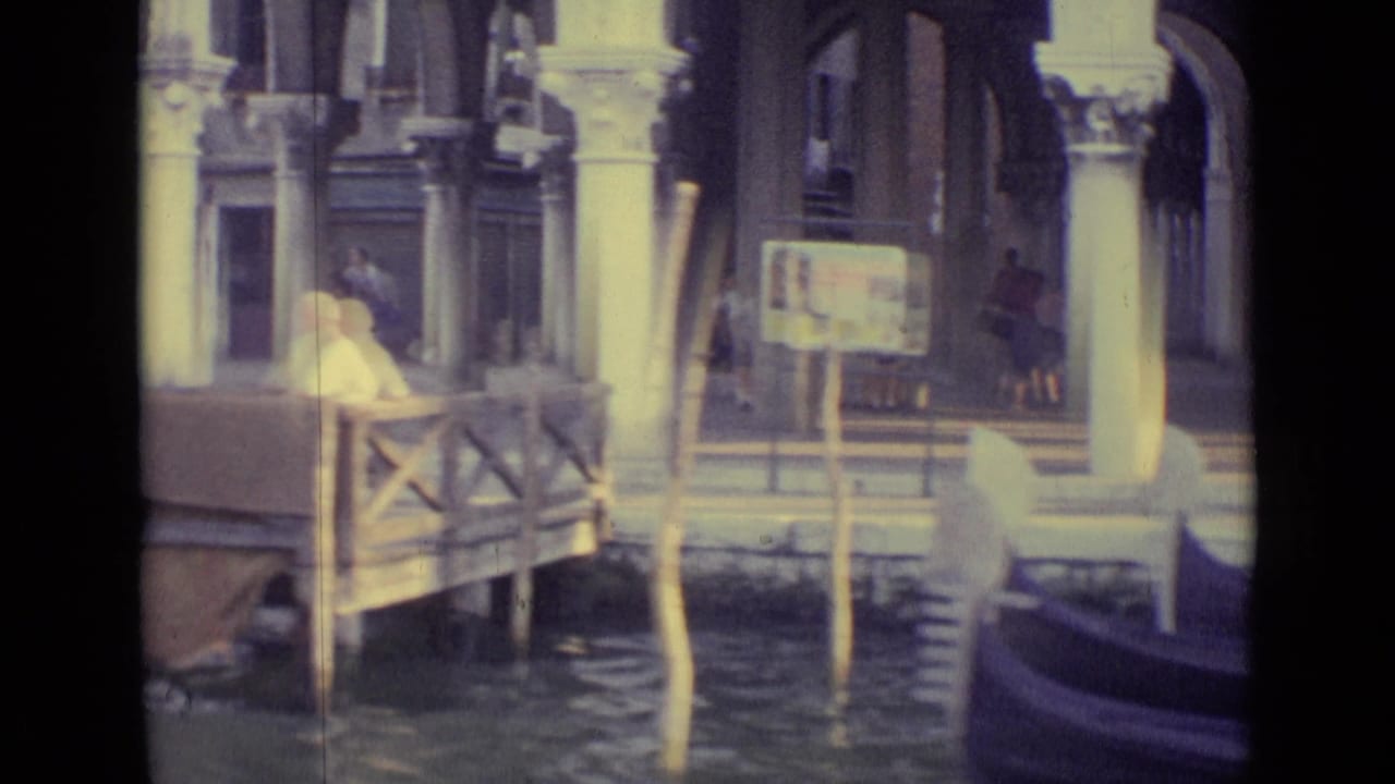 1962 Venice canal view with a small landing or bridge rail, mooring poles, water surface, columns, and a blurred gondola bow at the edge.
