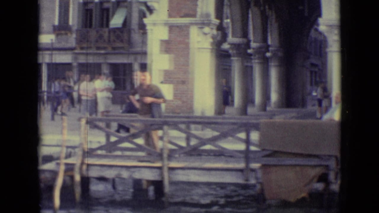 Venice canal-side scene from 1962, showing pedestrians near a wooden-railed bridge or landing, with columns and brick-and-stone buildings behind.
