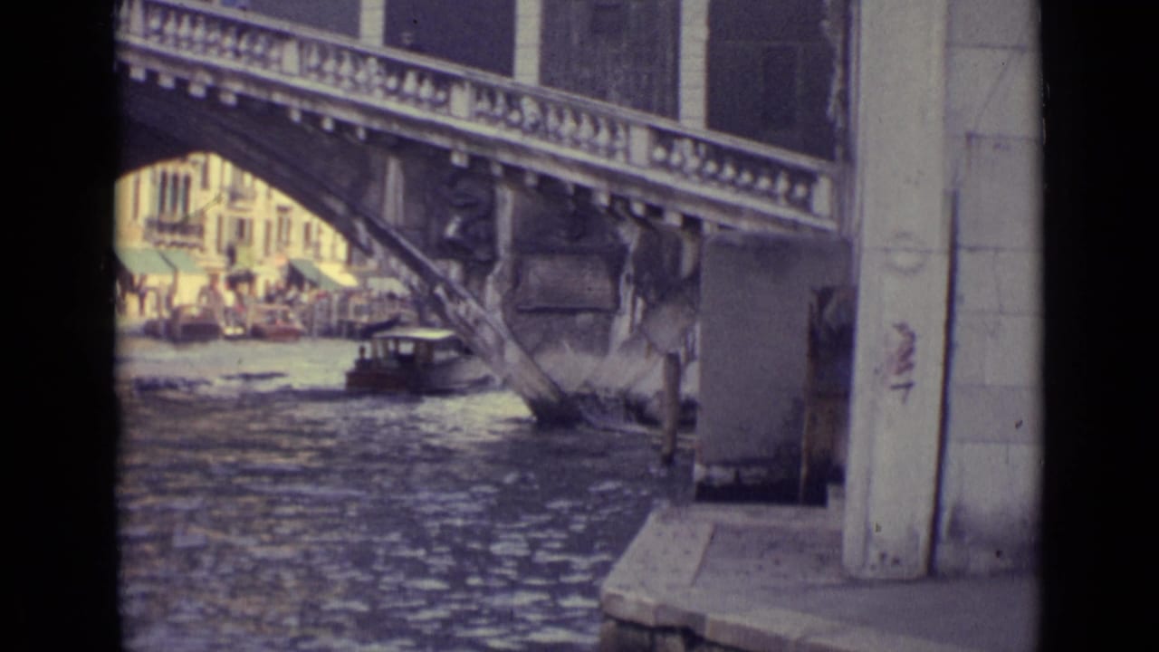 Canal view beneath a stone bridge with a small boat and waterfront buildings beyond.