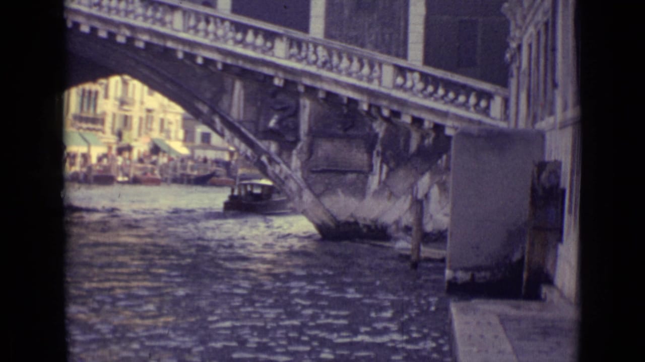 1962 Venice canal frame with rippling water, a stone bridge span, a small boat near the arch, and buildings visible through the opening.