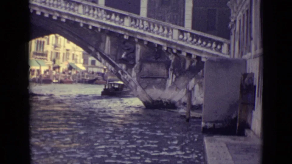 1962 Venice canal frame with rippling water, a stone bridge span, a small boat near the arch, and buildings visible through the opening.