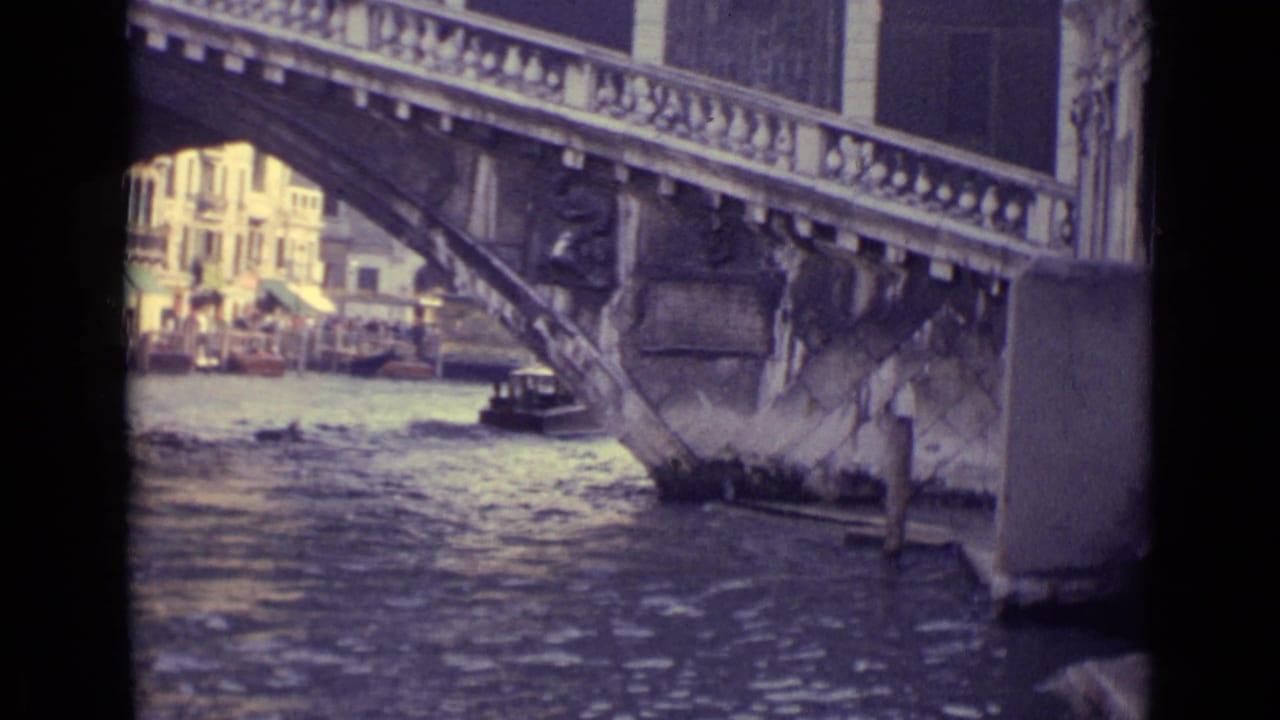 1962 Venice home-movie view of a stone bridge over canal water, with a small boat partly framed by the bridge arch and buildings beyond.