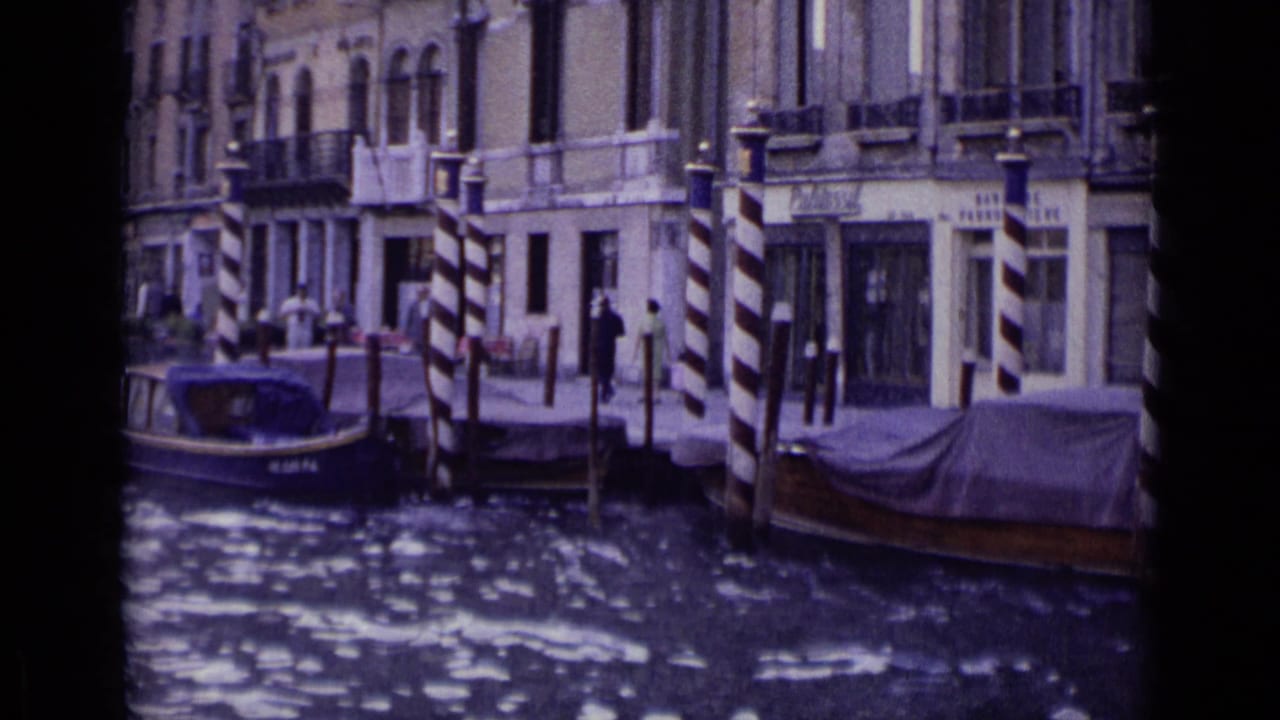 1962 Venice frame showing canal-front facades, striped mooring poles, moored boats, and distant pedestrians along the waterfront.