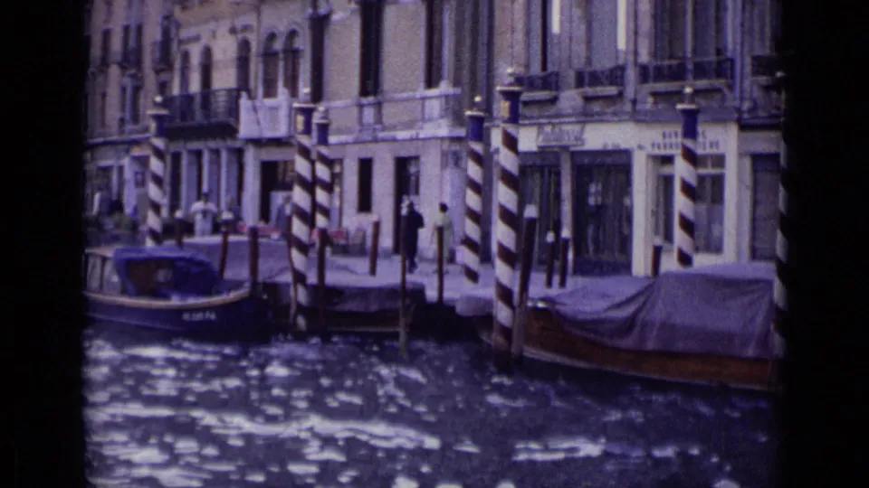 1962 Venice frame showing canal-front facades, striped mooring poles, moored boats, and distant pedestrians along the waterfront.