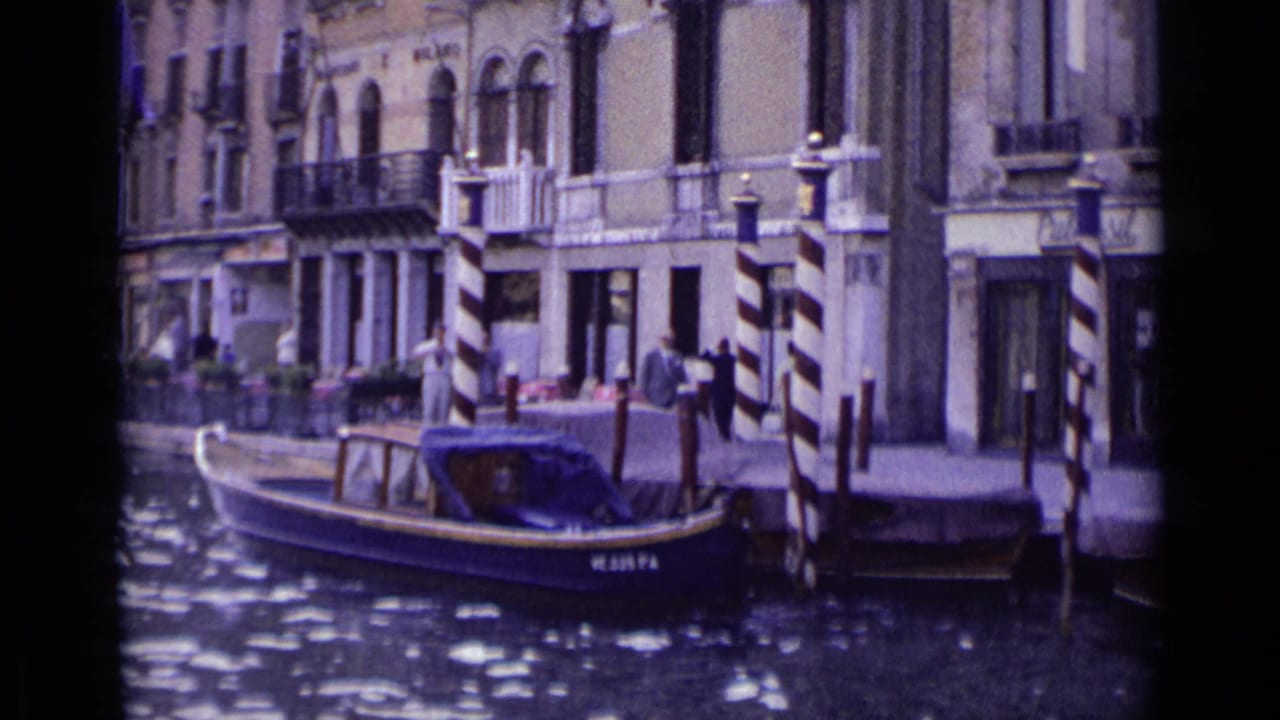 1962 Venice waterfront scene with a motorboat near striped mooring posts, arched building windows, balconies, and people on the canal edge.