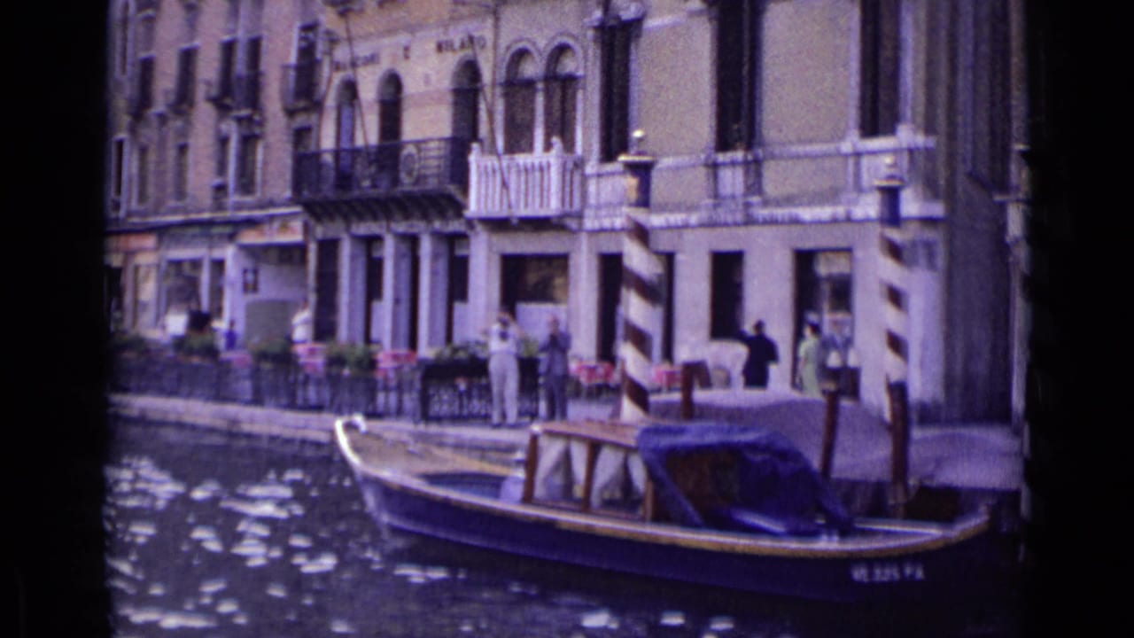 1962 Venice canal view showing a moored or passing motorboat, waterfront pedestrians, outdoor tables, balconies, and arched windows.