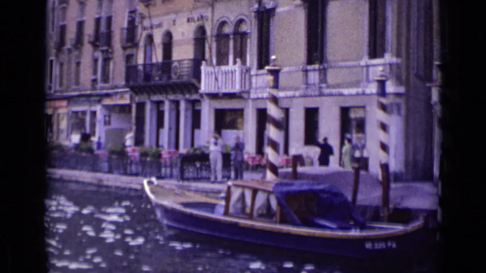 1962 Venice canal view showing a moored or passing motorboat, waterfront pedestrians, outdoor tables, balconies, and arched windows.