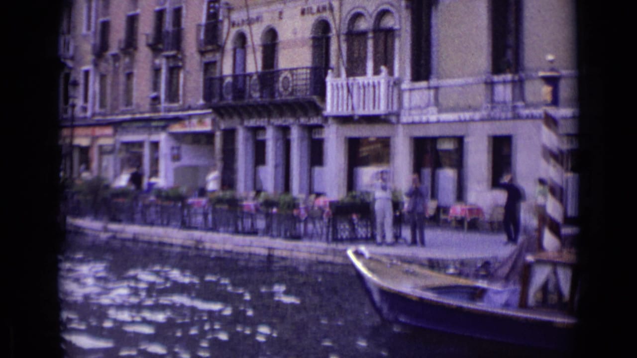 1962 Venice canal scene with people along the waterfront, tables near the building, striped mooring posts, and a small boat at the lower edge.