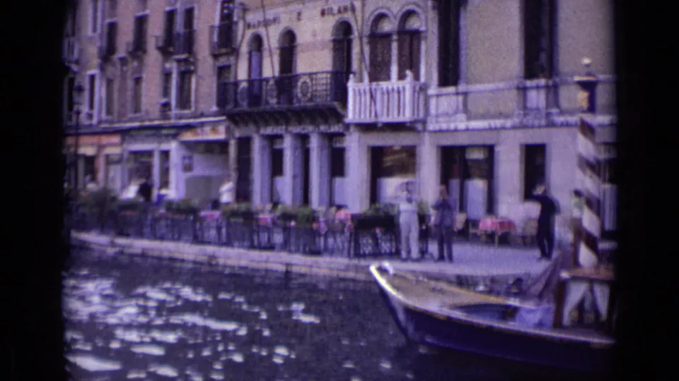 1962 Venice canal scene with people along the waterfront, tables near the building, striped mooring posts, and a small boat at the lower edge.