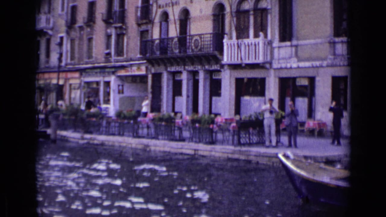 1962 Venice canal view from a moving gondola, showing waterfront shops, balconies, pedestrians, and cafe seating.