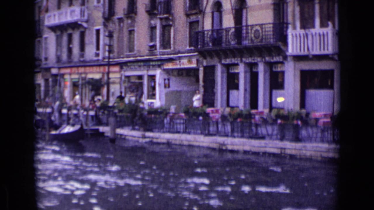 Waterfront shops and cafe tables with people along a canal, seen from across the water.