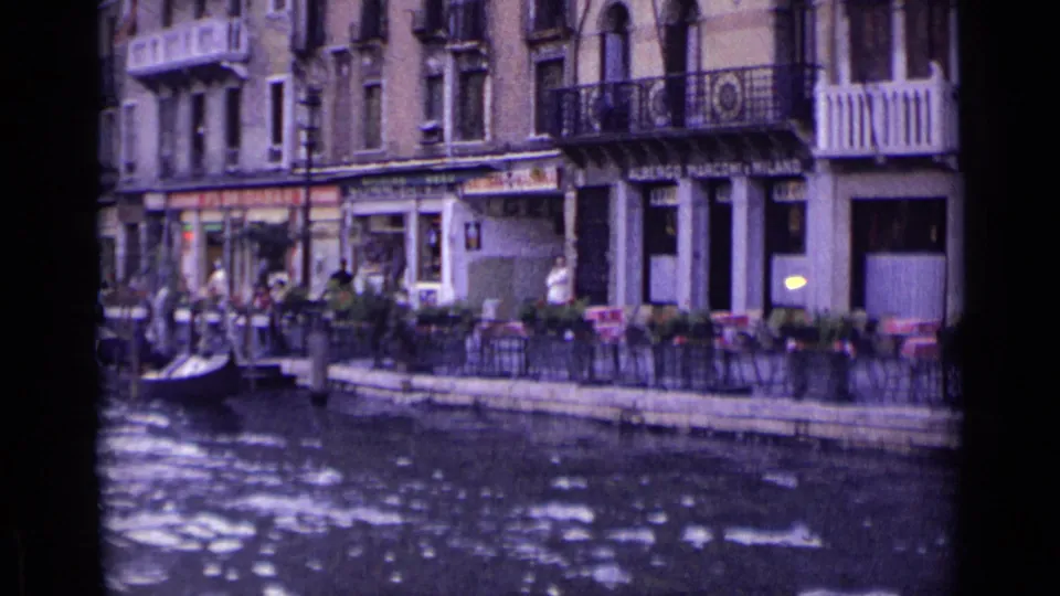 Venice canal frontage with pedestrians, seated visitors, shop signs, balconies, and a small boat near the sidewalk.