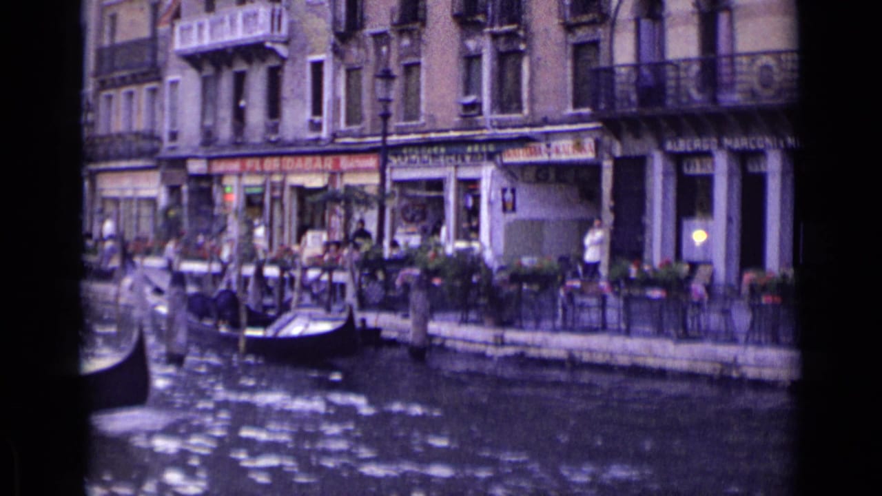 A gondolier moves through a Venice canal near visible shop signs, outdoor tables, balconies, and people on the sidewalk.