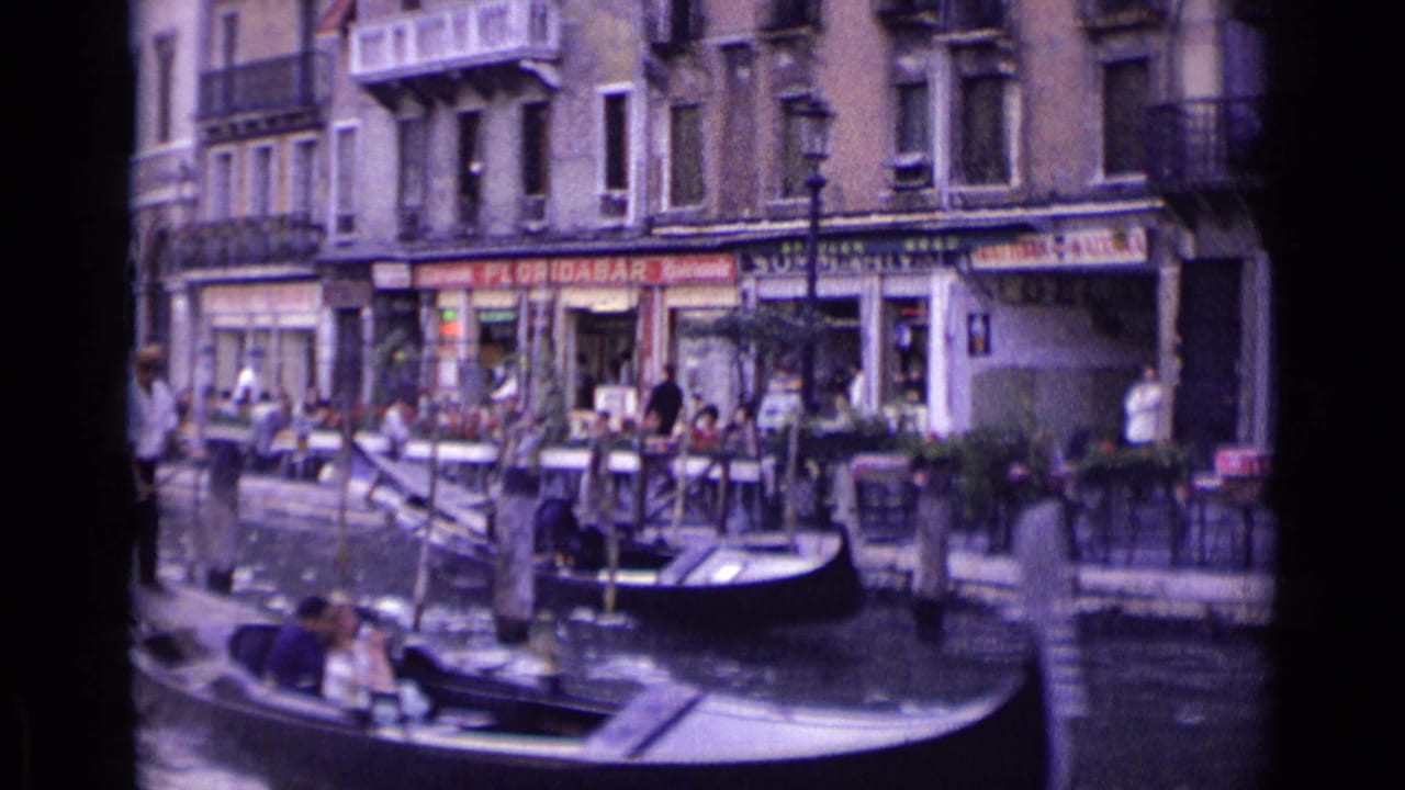 1962 Venice scene with gondolas in the foreground and canal-side shops, cafe tables, pedestrians, and balconies behind them.