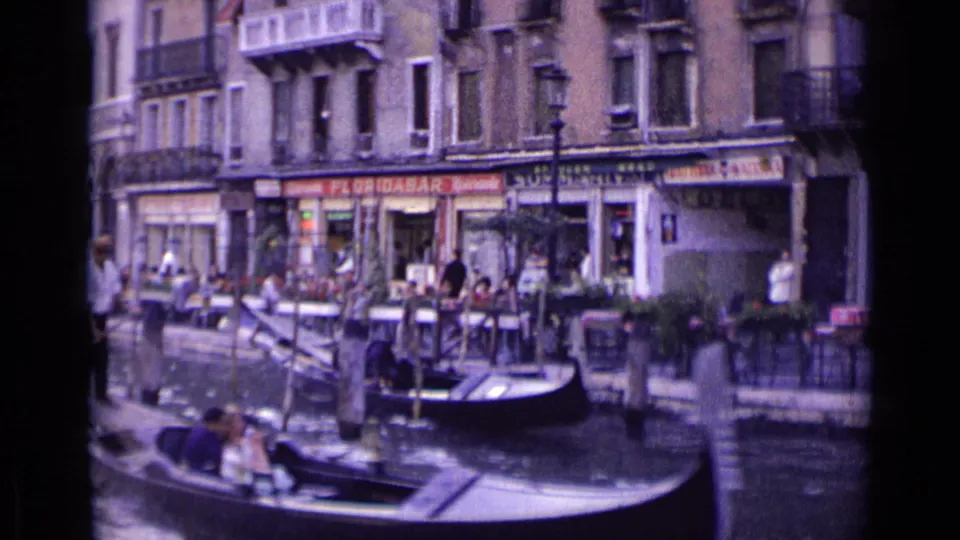 1962 Venice scene with gondolas in the foreground and canal-side shops, cafe tables, pedestrians, and balconies behind them.