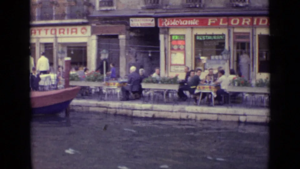 1962 Venice canal-side restaurant view with seated cafe patrons, red boat edge, storefront signs, and water in the foreground.