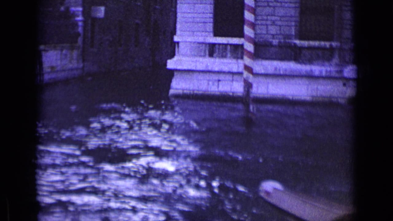 Canal water beside stone buildings and striped mooring posts, with part of a boat at the lower edge.