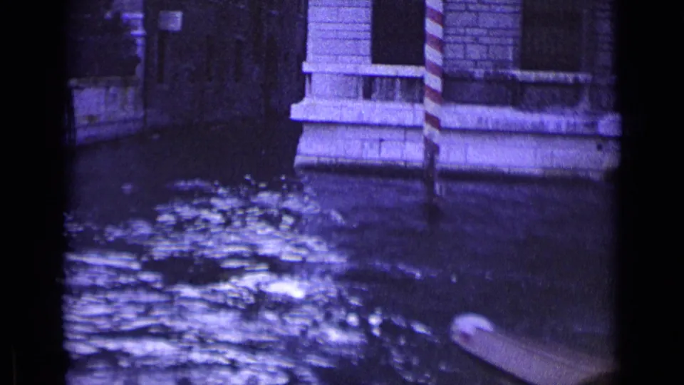 1962 Venice canal view with rippling water, stone facades, striped mooring posts, and the edge of a small boat in frame.