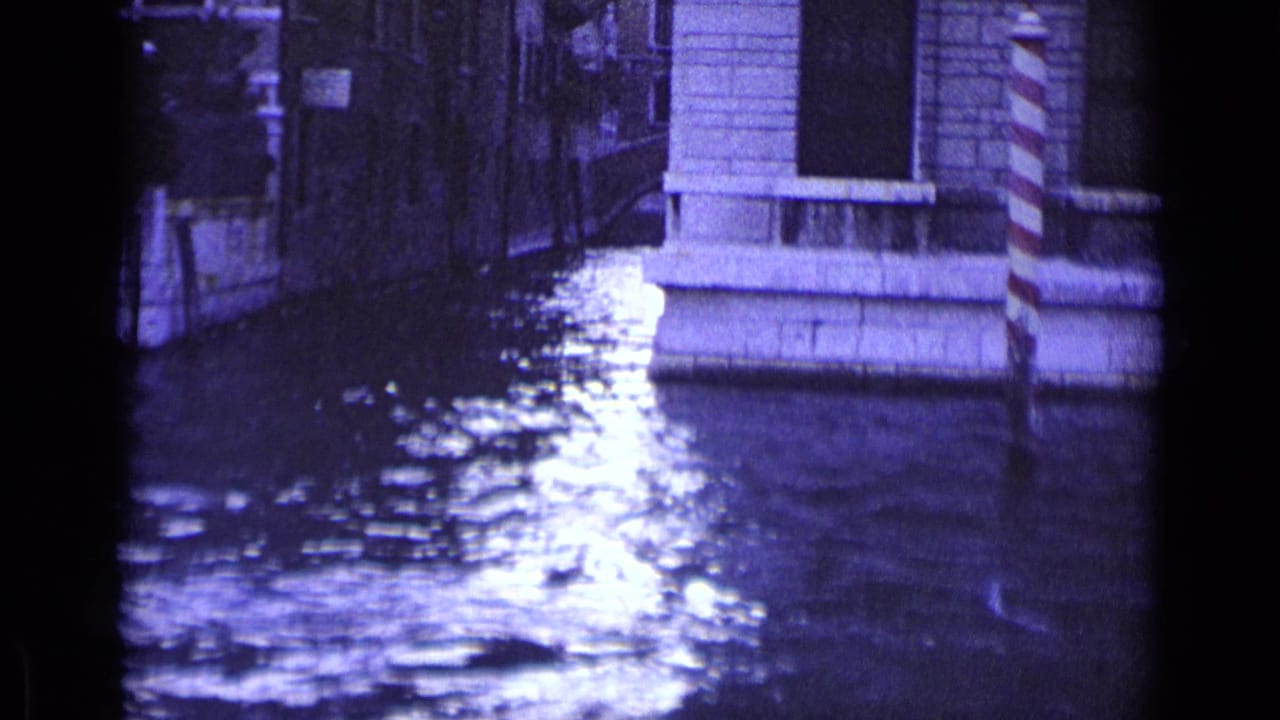 Canal water reflecting light between stone buildings and striped mooring posts.