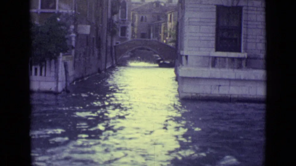 1962 Venice view down a narrow canal toward a small arched bridge, with water reflecting pale light between buildings.