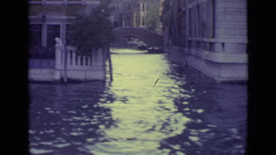 In 1962 Venice, a man appears to row a small boat along a narrow canal bordered by stone buildings and a small arched bridge.