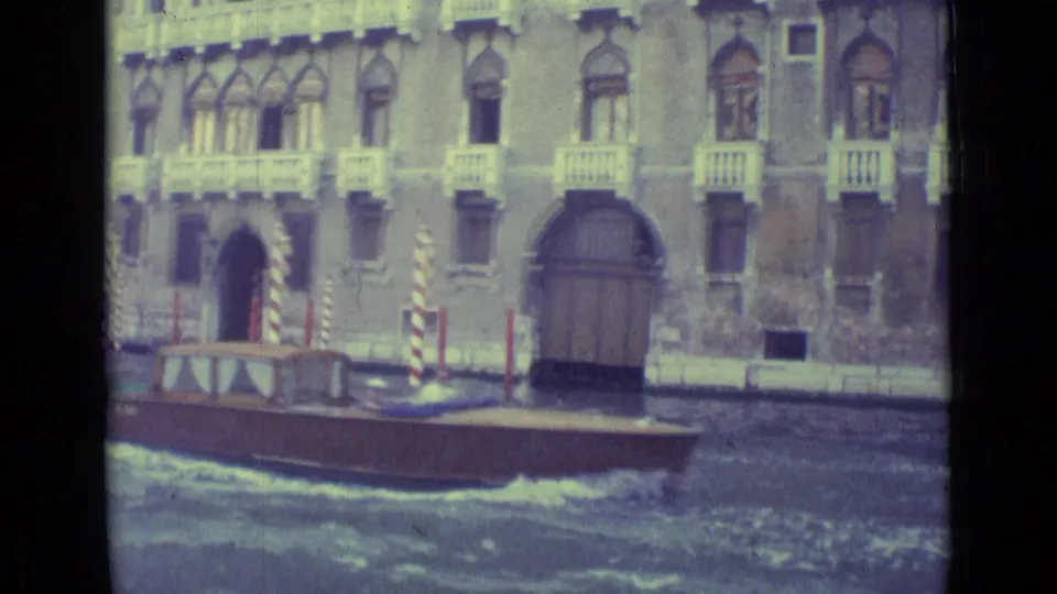 1962 Venice 8mm frame showing a wooden canal boat moving past arched doors, balconies, and striped mooring poles.