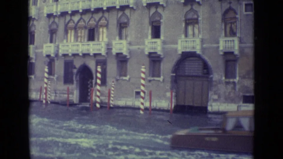 1962 Venice canal view with ornate windows, arched entry, striped poles, and a wooden boat partly visible at right.