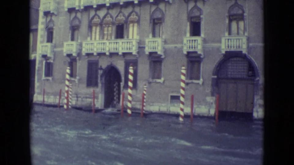 1962 Venice 8mm frame of a canal-facing palazzo with balconies, dark arched doorway, striped poles, and rippled water.