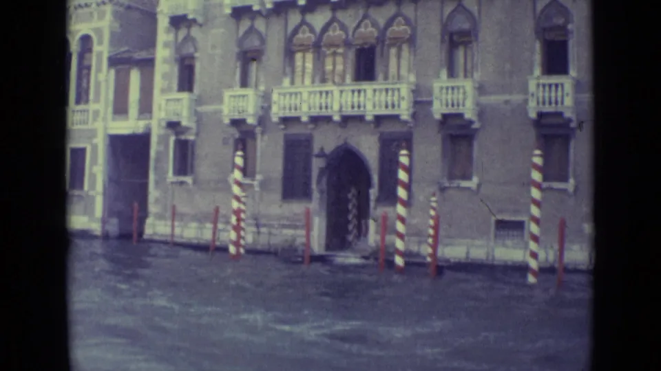 1962 Venice home-movie frame showing a palazzo exterior, striped mooring poles, windows, and canal water from a moving boat.