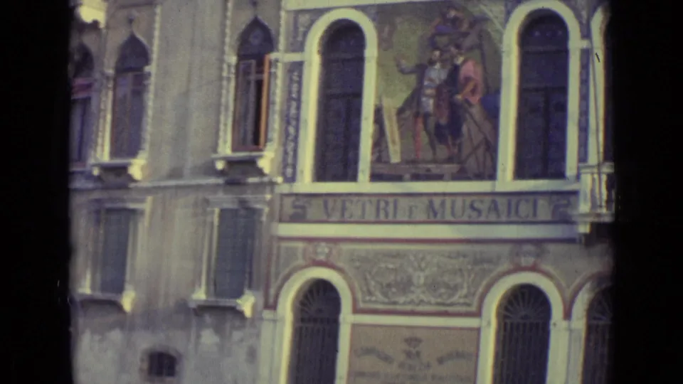 Final 1962 Venice frame showing a Vetri e Musaici sign under painted figures, flanked by arched windows and barred ground-floor openings.