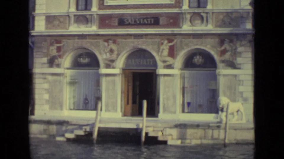 1962 Venice 8mm frame showing the SALVIATI waterfront facade, arched entrances, canal posts, steps, and a white lion statue.