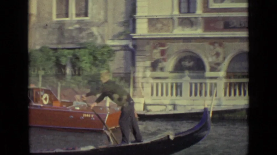 1962 Venice 8mm frame with a gondolier in dark clothing passing an ornate canal facade and a wooden motorboat.