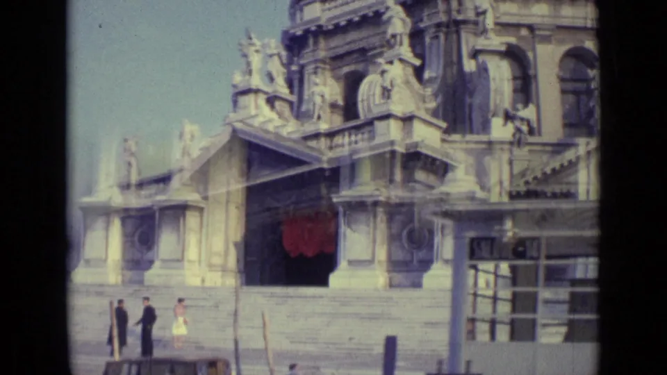 1962 Venice 8mm frame showing a domed historic facade with statues, a red doorway covering, canal posts, and small figures near the entrance.