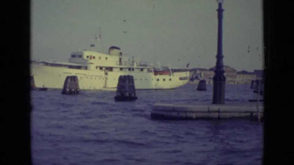 A white passenger ship moves across the Venice lagoon near wooden mooring posts, seen through a dark boat-window frame.