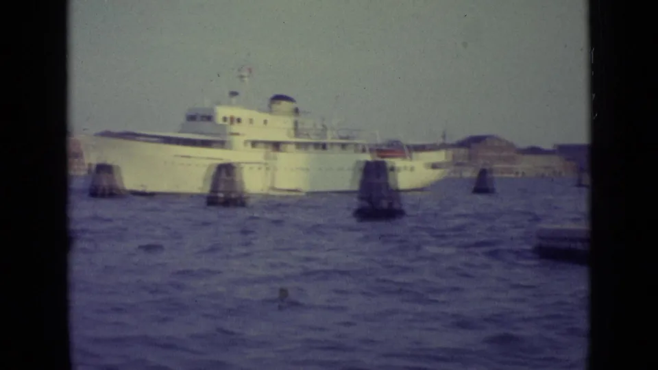The white passenger ship continues across the Venice lagoon, partially framed by the filming boat and wooden navigation posts.