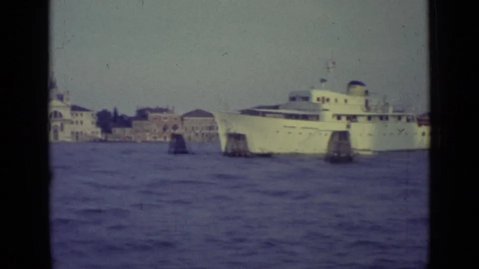 A white passenger ship passes near Venice shoreline buildings, with lagoon posts and open water in the foreground.