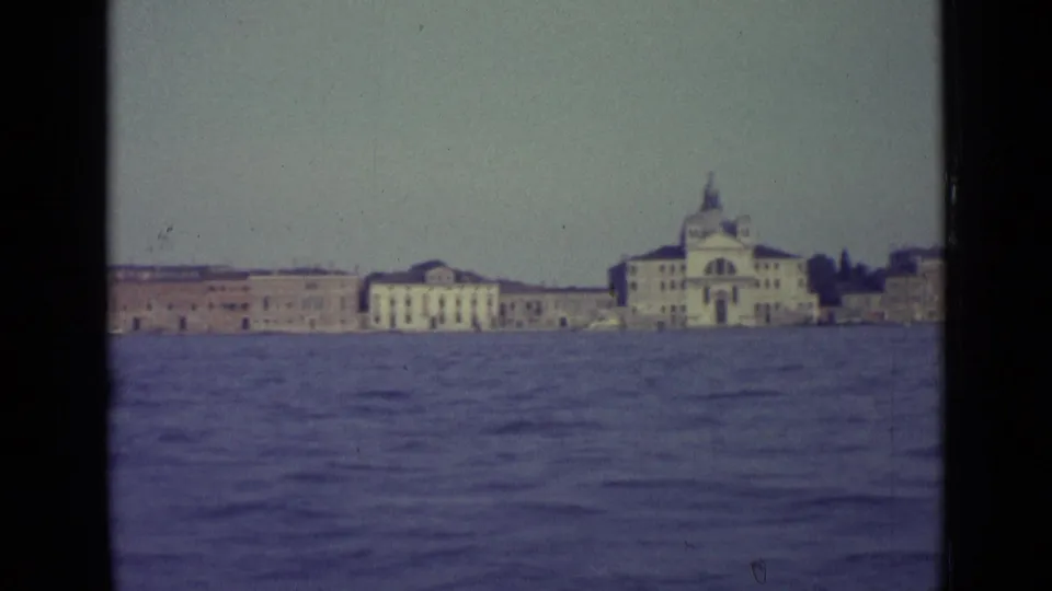 1962 Venice 8mm frame looking across water toward historic waterfront buildings and a domed church-like landmark.