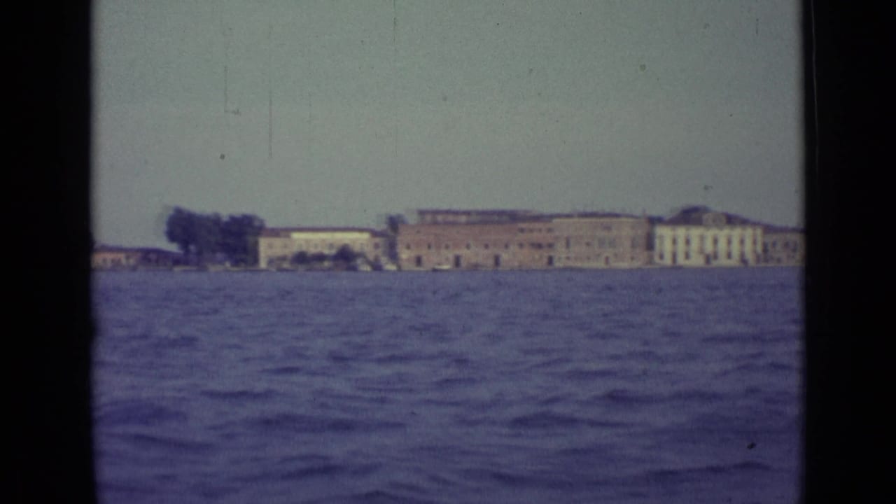 1962 Venice waterfront seen across choppy canal water, with distant low buildings and dark 8mm frame edges.