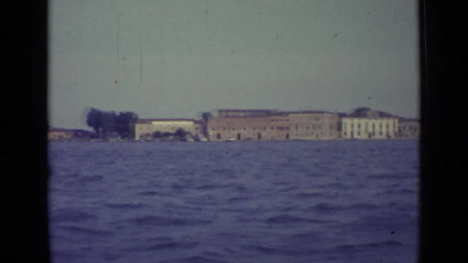1962 Venice waterfront seen across choppy canal water, with distant low buildings and dark 8mm frame edges.