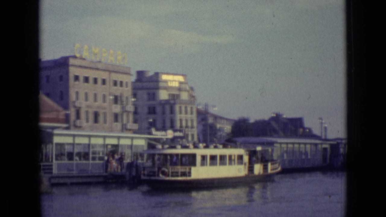 A Venice dock area in 1962 with a small boat at the landing, people nearby, waterfront buildings, and a rooftop Campari sign.