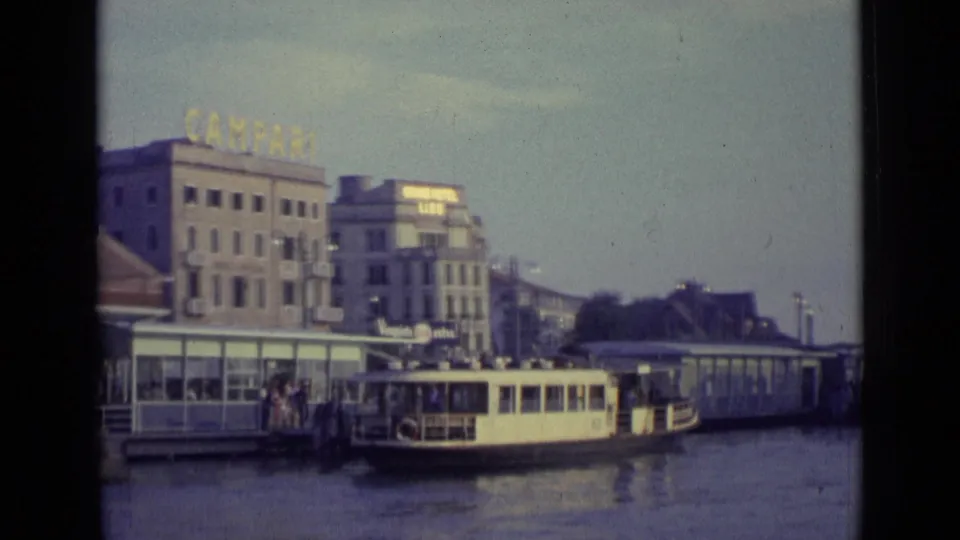 A Venice dock area in 1962 with a small boat at the landing, people nearby, waterfront buildings, and a rooftop Campari sign.