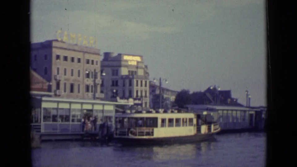 1962 Venice waterfront landing with a cream-colored boat, gathered people, dock structures, and Campari signage on the building above.